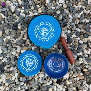 May include: Three blue singing bowls with a white design of a chakra symbol. The bowls are on a bed of small gray and white stones. A wooden striker is included.