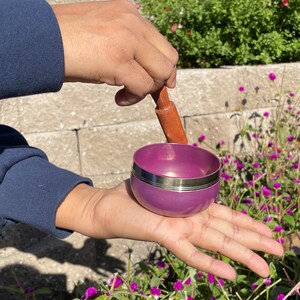 May include: A purple singing bowl with a silver rim and a wooden striker. The bowl is being held in a person's hand.