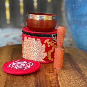 Puede incluir: Un cuenco cantor rojo con un borde plateado, un mazo de madera y una bolsa de terciopelo rojo con bordados dorados. El cuenco cantor est&aacute; sobre una superficie de madera.