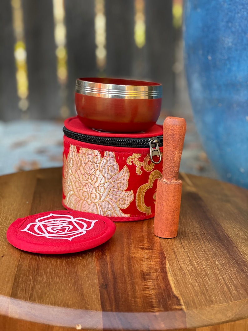 Puede incluir: Un cuenco cantor rojo con un borde plateado, un mazo de madera y una bolsa de terciopelo rojo con bordados dorados. El cuenco cantor est&aacute; sobre una superficie de madera.