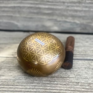Puede incluir: Un cuenco cantor dorado con un dise&ntilde;o de flor de la vida y las palabras "Made in Nepal" en el lateral. El cuenco est&aacute; sobre una superficie de madera junto a un mazo de madera.