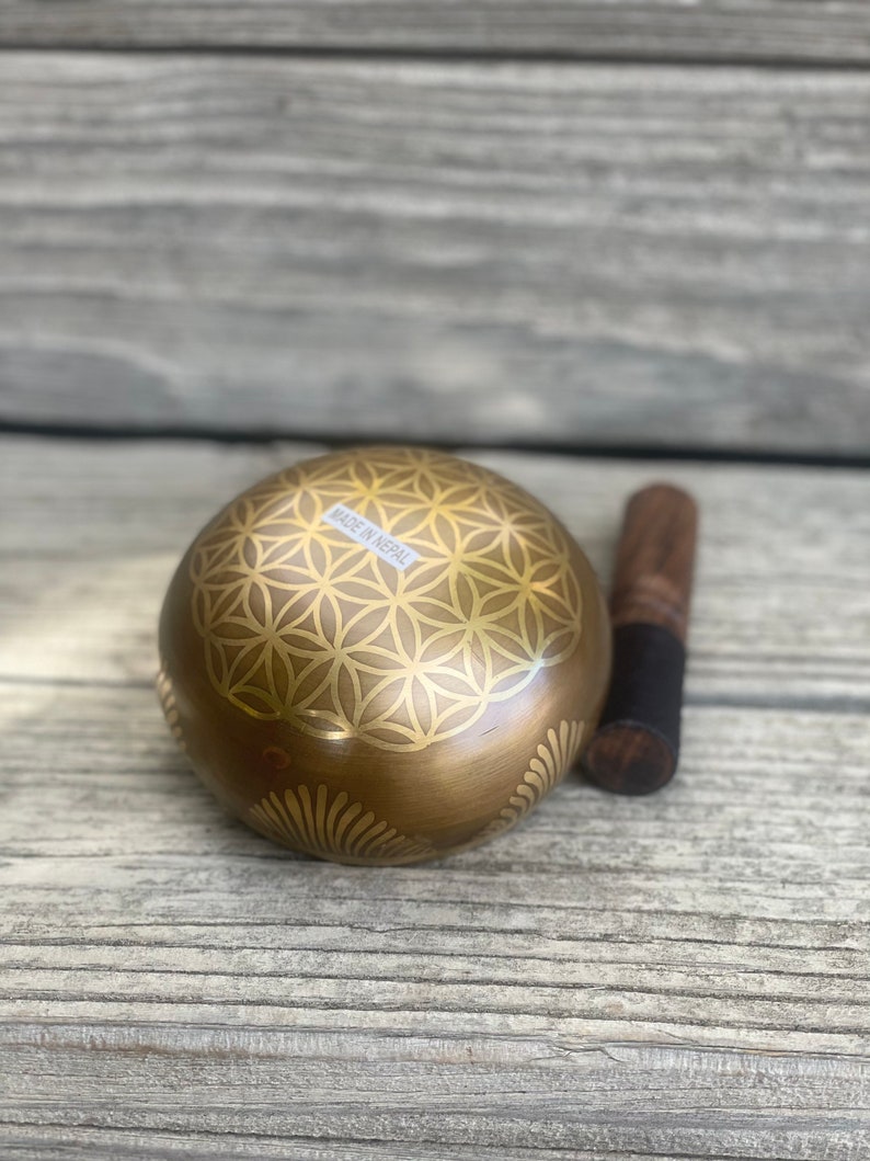 Puede incluir: Un cuenco cantor dorado con un dise&ntilde;o de flor de la vida y las palabras "Made in Nepal" en el lateral. El cuenco est&aacute; sobre una superficie de madera junto a un mazo de madera.