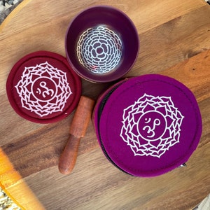 May include: Two purple singing bowls with a white mandala design and the Sanskrit symbol Om. One bowl is in a purple pouch with a white mandala design and the Sanskrit symbol Om. A wooden striker is included.