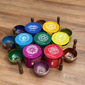 May include: Set of seven singing bowls with wooden strikers, each bowl is a different color and has a different chakra symbol and name printed on it. The bowls are arranged in a circle on a wooden surface.