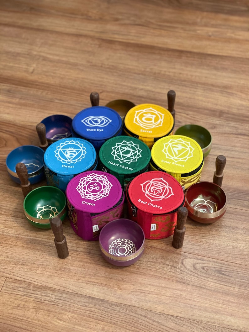 May include: Set of seven singing bowls with wooden strikers, each bowl is a different color and has a different chakra symbol and name printed on it. The bowls are arranged in a circle on a wooden surface.