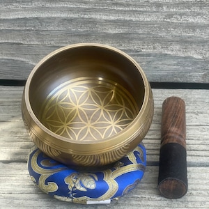 Puede incluir: Un cuenco cantor dorado con un dise&ntilde;o de flor de la vida, un coj&iacute;n azul y dorado y un mazo de madera con una punta de goma negra.