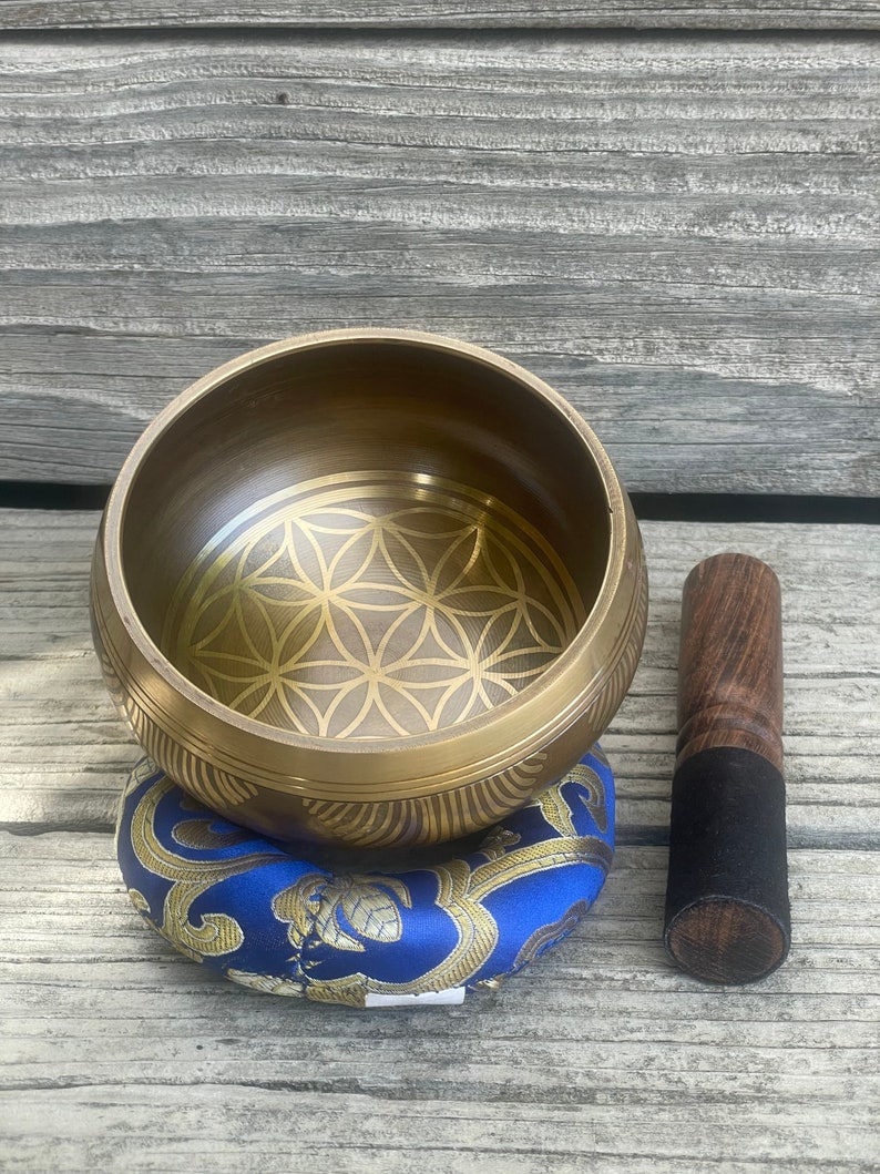 Puede incluir: Un cuenco cantor dorado con un dise&ntilde;o de flor de la vida, un coj&iacute;n azul y dorado y un mazo de madera con una punta de goma negra.