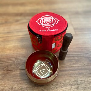 May include: Red singing bowl with a gold rim and a white root chakra symbol. The bowl comes with a wooden striker and a red fabric carrying case with a zipper closure and the words "Root Chakra" printed on it.