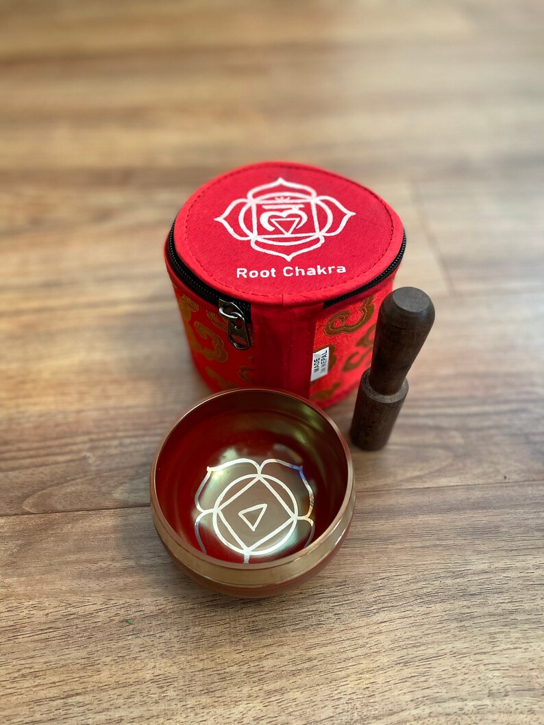 May include: Red singing bowl with a gold rim and a white root chakra symbol. The bowl comes with a wooden striker and a red fabric carrying case with a zipper closure and the words "Root Chakra" printed on it.