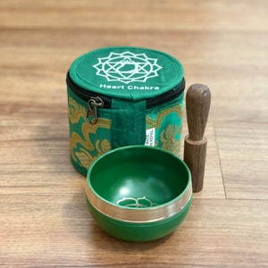 May include: Green singing bowl with a silver rim and a wooden striker. The bowl is in a green and gold fabric pouch with the words "Heart Chakra" printed on it. The pouch has a zipper closure and a silver pull tab. The pouch is labeled "Made in Nepal".