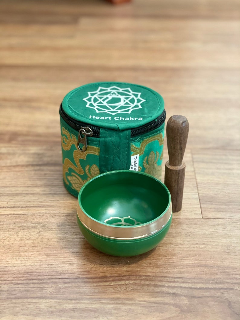 May include: Green singing bowl with a silver rim and a wooden striker. The bowl is in a green and gold fabric pouch with the words "Heart Chakra" printed on it. The pouch has a zipper closure and a silver pull tab. The pouch is labeled "Made in Nepal".
