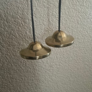 May include: Two small, round, brass bells hanging from black cords. The bells are slightly different sizes and have a smooth, shiny finish.