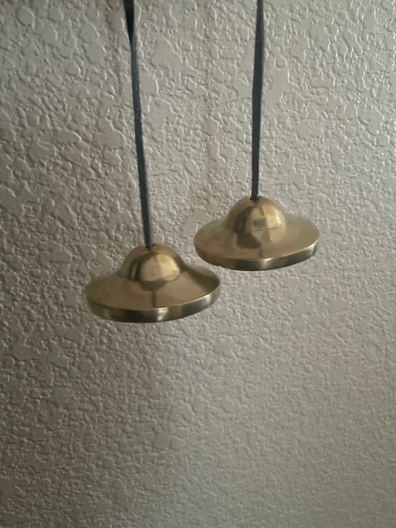 May include: Two small, round, brass bells hanging from black cords. The bells are slightly different sizes and have a smooth, shiny finish.