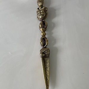 May include: A gold-colored metal dagger with intricate carvings and a series of decorative beads. The dagger has a pointed tip and a small, detailed head at the top.