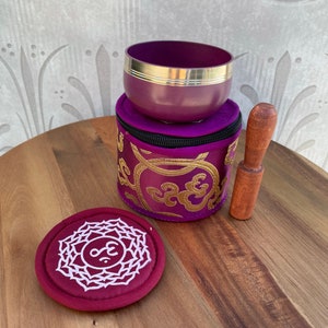 May include: A purple singing bowl with a gold rim and a wooden striker. The bowl is in a purple and gold embroidered pouch with a zipper. A red round cushion with a white symbol is also included.