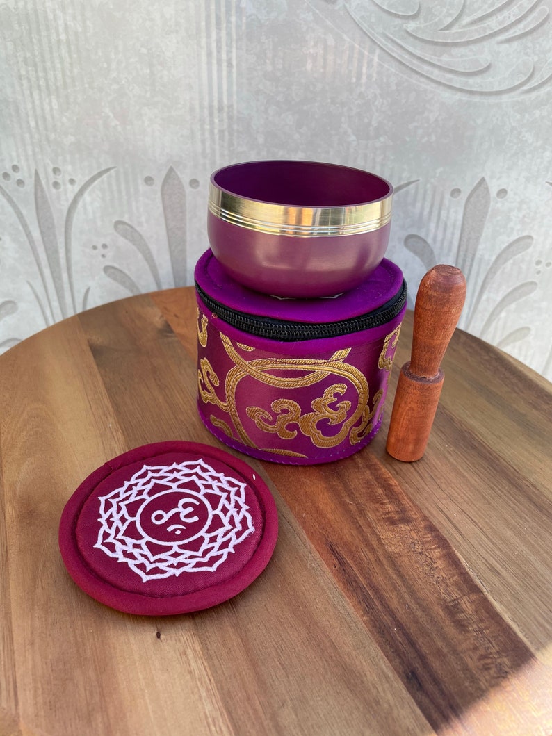 May include: A purple singing bowl with a gold rim and a wooden striker. The bowl is in a purple and gold embroidered pouch with a zipper. A red round cushion with a white symbol is also included.