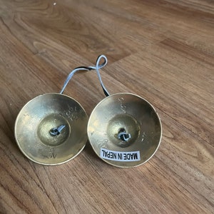 May include: Two small, round, brass cymbals connected by a black cord. Each cymbal has a small, silver bell in the centre. The cymbals are labelled "MADE IN NEPAL".