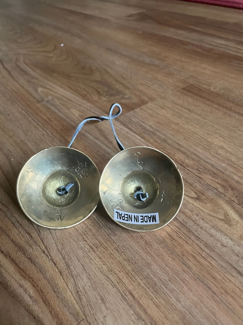 May include: Two small, round, brass cymbals connected by a black cord. Each cymbal has a small, silver bell in the centre. The cymbals are labelled "MADE IN NEPAL".