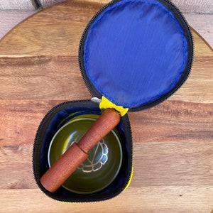 May include: A yellow and black zippered pouch with a blue lining. The pouch contains a green singing bowl and a wooden striker.