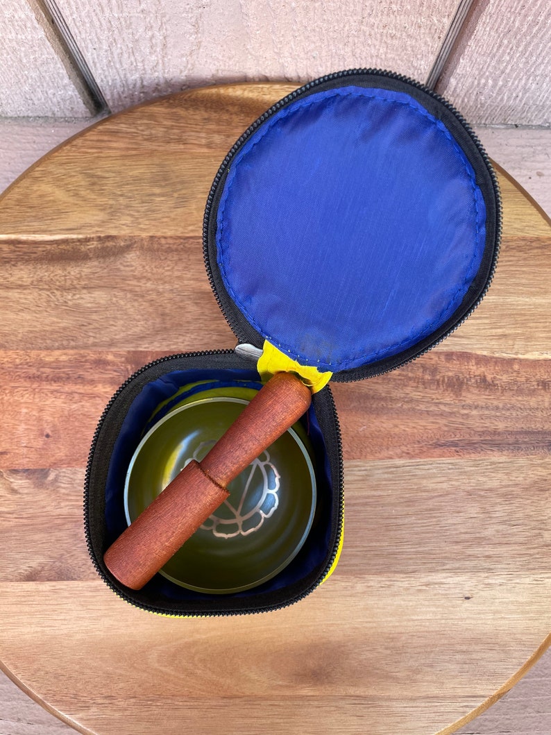May include: A yellow and black zippered pouch with a blue lining. The pouch contains a green singing bowl and a wooden striker.