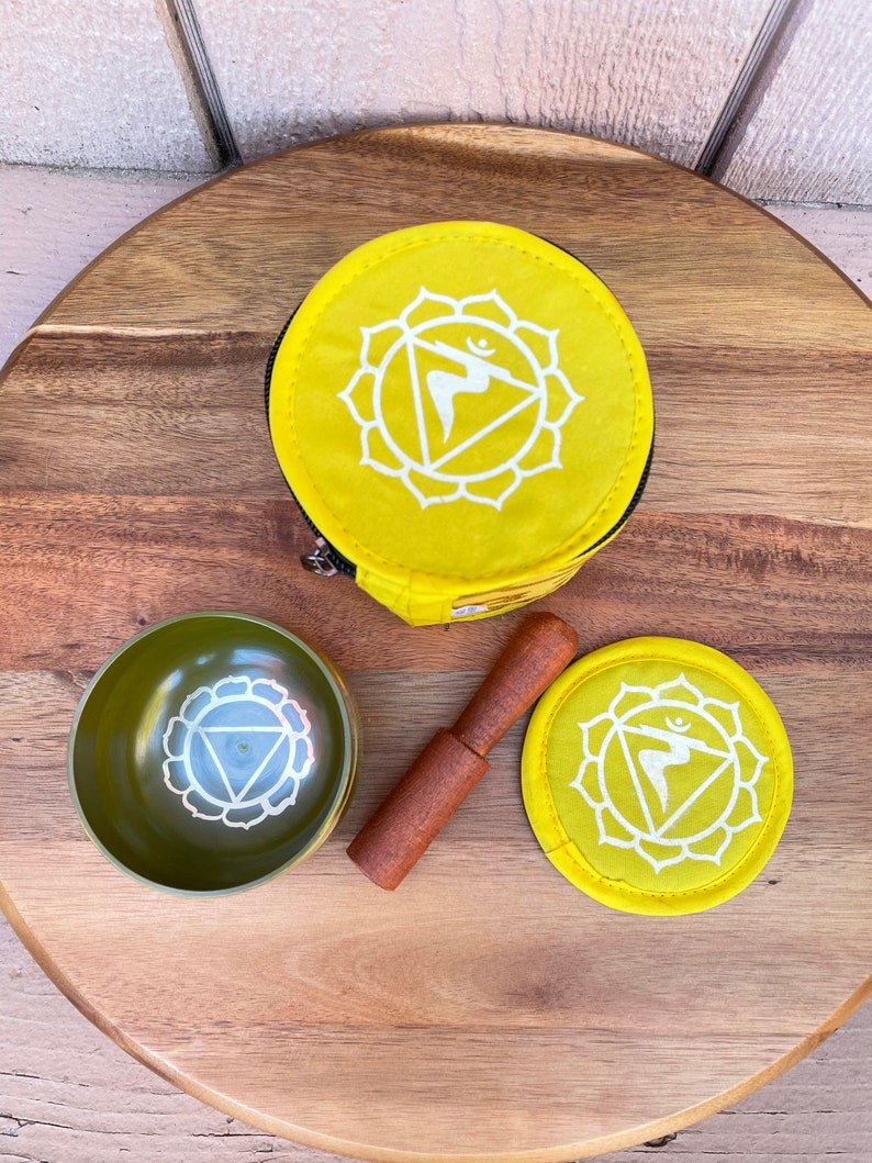 May include: A yellow singing bowl set with a wooden striker, a yellow pouch, and a yellow cushion. The bowl and cushion feature a white symbol of the solar plexus chakra.