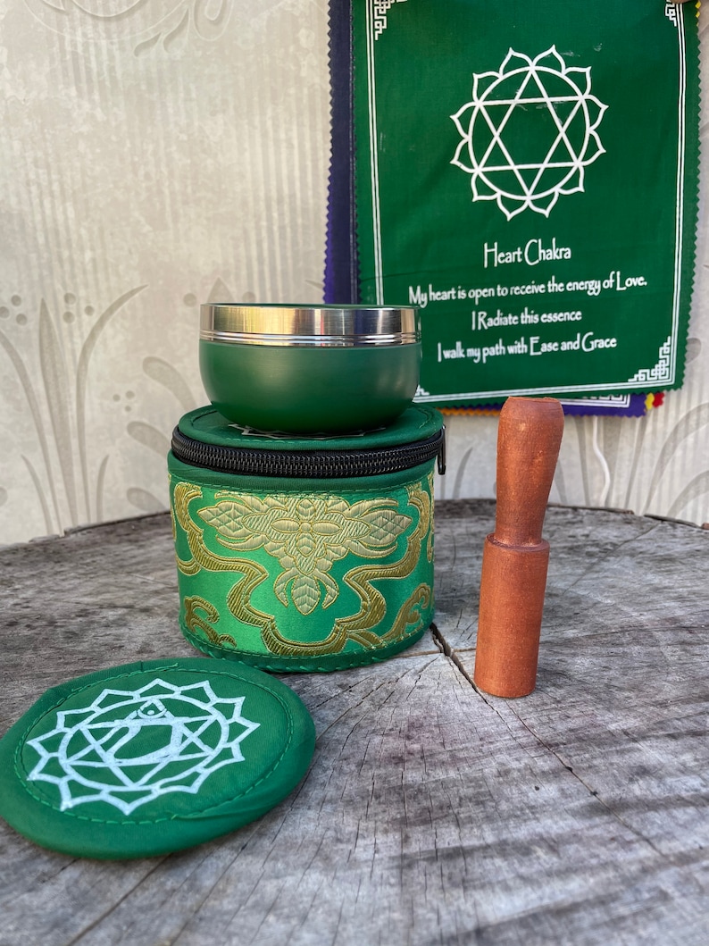May include: Green singing bowl set with a wooden striker, a green cushion, and a green pouch with gold embroidery. The pouch has a zipper closure. The singing bowl is made of metal and has a silver rim. The set also includes a green fabric with a white heart chakra symbol and the text "Heart Chakra" in white. The text below the symbol reads "My heart is open to receive the energy of Love. I Radiate this essence I walk my path with Ease and Grace."