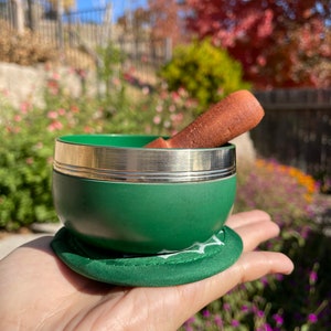 May include: A green singing bowl with a silver rim and a brown wooden striker. The bowl is sitting on a green cushion.