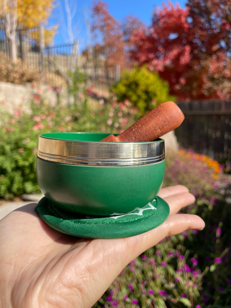May include: A green singing bowl with a silver rim and a brown wooden striker. The bowl is sitting on a green cushion.