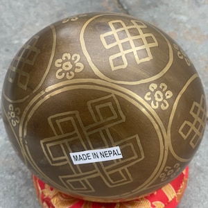 Puede incluir: Un cuenco cantor marr&oacute;n con intrincados dise&ntilde;os dorados y el texto "MADE IN NEPAL". El cuenco est&aacute; sentado sobre un coj&iacute;n rojo con dise&ntilde;os dorados.