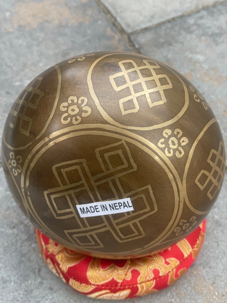 Puede incluir: Un cuenco cantor marr&oacute;n con intrincados dise&ntilde;os dorados y el texto "MADE IN NEPAL". El cuenco est&aacute; sentado sobre un coj&iacute;n rojo con dise&ntilde;os dorados.