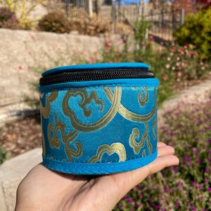 May include: A small, round, blue and gold patterned storage box with a zipper closure. The box is made of fabric and has a soft, plush texture.
