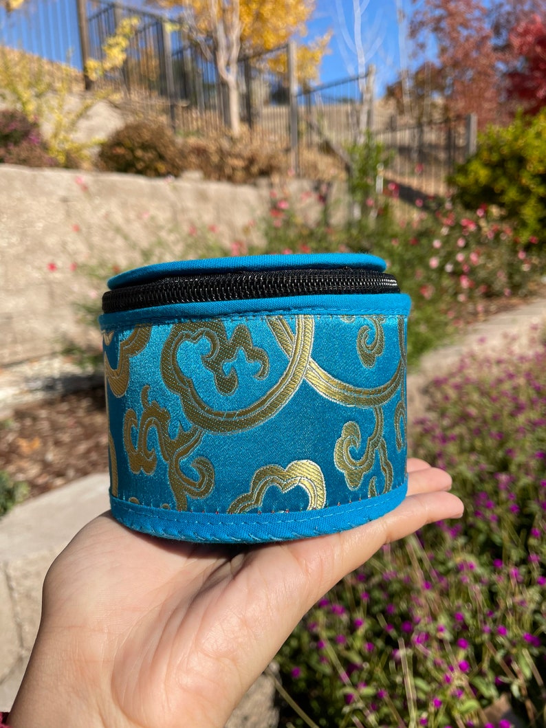 May include: A small, round, blue and gold patterned storage box with a zipper closure. The box is made of fabric and has a soft, plush texture.