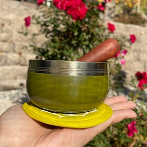 May include: A green singing bowl with a silver rim and a wooden striker. The bowl is sitting on a yellow cushion.