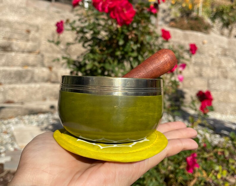May include: A green singing bowl with a silver rim and a wooden striker. The bowl is sitting on a yellow cushion.