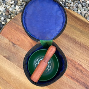 May include: A green singing bowl with a silver lotus design, a wooden striker, and a black zippered carrying case.