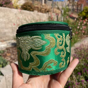 May include: A small, round, green zippered pouch with a gold and white floral pattern. The pouch is made of a soft, silky fabric.