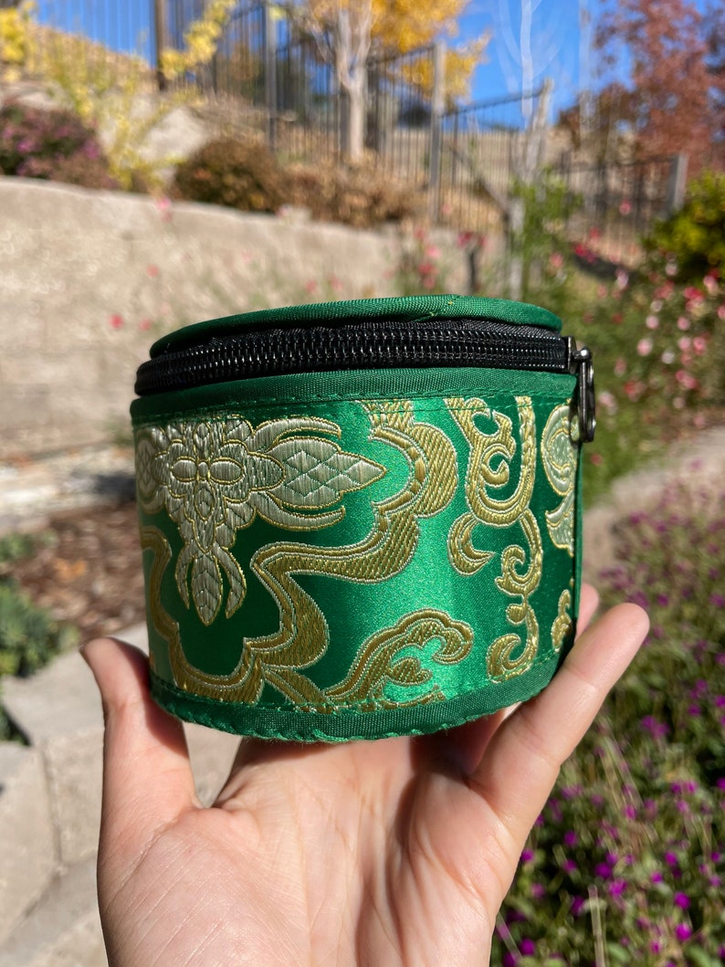 May include: A small, round, green zippered pouch with a gold and white floral pattern. The pouch is made of a soft, silky fabric.