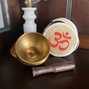 May include: A gold singing bowl with a wooden striker, a beige fabric pouch with a red Om symbol, and a white marble base.
