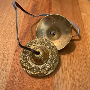 May include: Two small, round, gold-coloured cymbals with intricate designs. The cymbals are connected by a black leather strap.