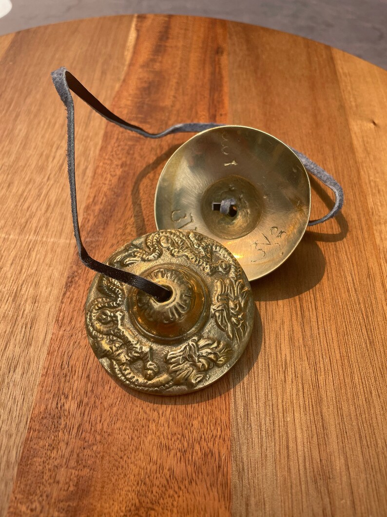 May include: Two small, round, gold-coloured cymbals with intricate designs. The cymbals are connected by a black leather strap.