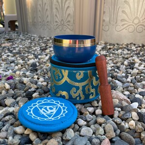 May include: A blue singing bowl with a gold rim and a wooden striker. The bowl is in a blue and gold patterned pouch. There is a blue round cushion with a white symbol on it.