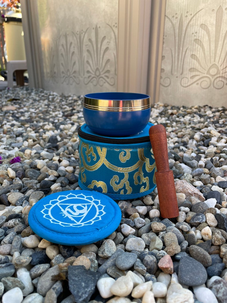 May include: A blue singing bowl with a gold rim and a wooden striker. The bowl is in a blue and gold patterned pouch. There is a blue round cushion with a white symbol on it.