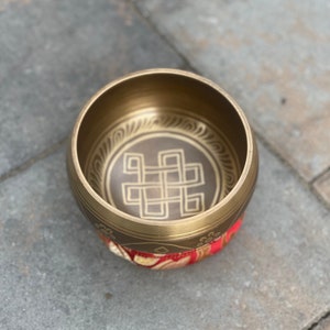 Puede incluir: Un cuenco de canto de lat&oacute;n con un dise&ntilde;o decorativo de un nudo dentro de un c&iacute;rculo. El cuenco est&aacute; apoyado en un coj&iacute;n rojo y dorado.