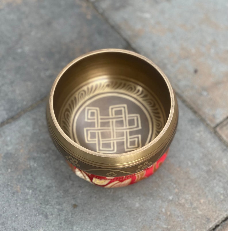 Puede incluir: Un cuenco de canto de lat&oacute;n con un dise&ntilde;o decorativo de un nudo dentro de un c&iacute;rculo. El cuenco est&aacute; apoyado en un coj&iacute;n rojo y dorado.