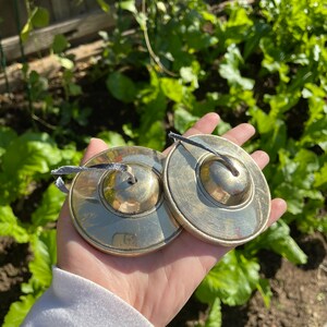 May include: Two small, round, brass cymbals with black string handles. The cymbals are held in a hand wearing a white long-sleeved shirt.