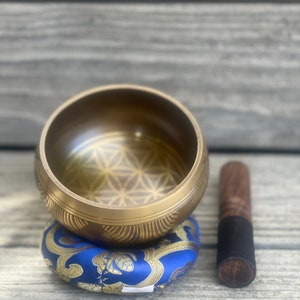 Puede incluir: Un cuenco cantor dorado con un dise&ntilde;o de flor de la vida se sienta sobre un coj&iacute;n azul y dorado con estampado. Un mazo de madera con una punta de goma negra est&aacute; al lado del cuenco.