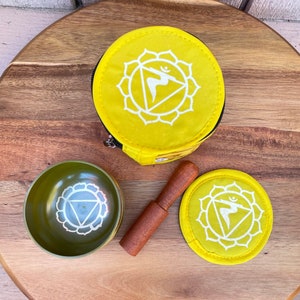 Puede incluir: Un juego de cuencos cantores de meditaci&oacute;n amarillo con un mazo de madera, una bolsa de tela amarilla y un coj&iacute;n de tela amarilla. El cuenco y el coj&iacute;n presentan un s&iacute;mbolo blanco del chakra del plexo solar.