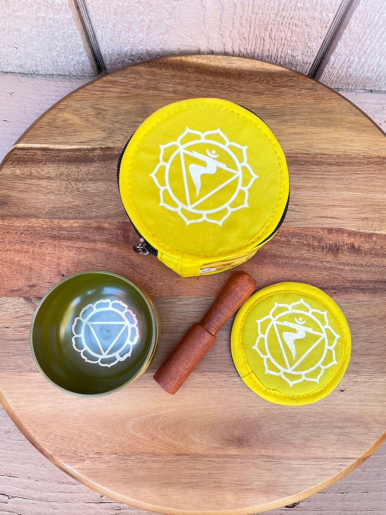 Puede incluir: Un juego de cuencos cantores de meditaci&oacute;n amarillo con un mazo de madera, una bolsa de tela amarilla y un coj&iacute;n de tela amarilla. El cuenco y el coj&iacute;n presentan un s&iacute;mbolo blanco del chakra del plexo solar.