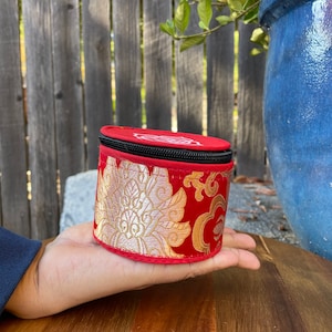 May include: A small, round, red and gold fabric box with a black zipper. The box has a floral pattern and is being held in a hand.