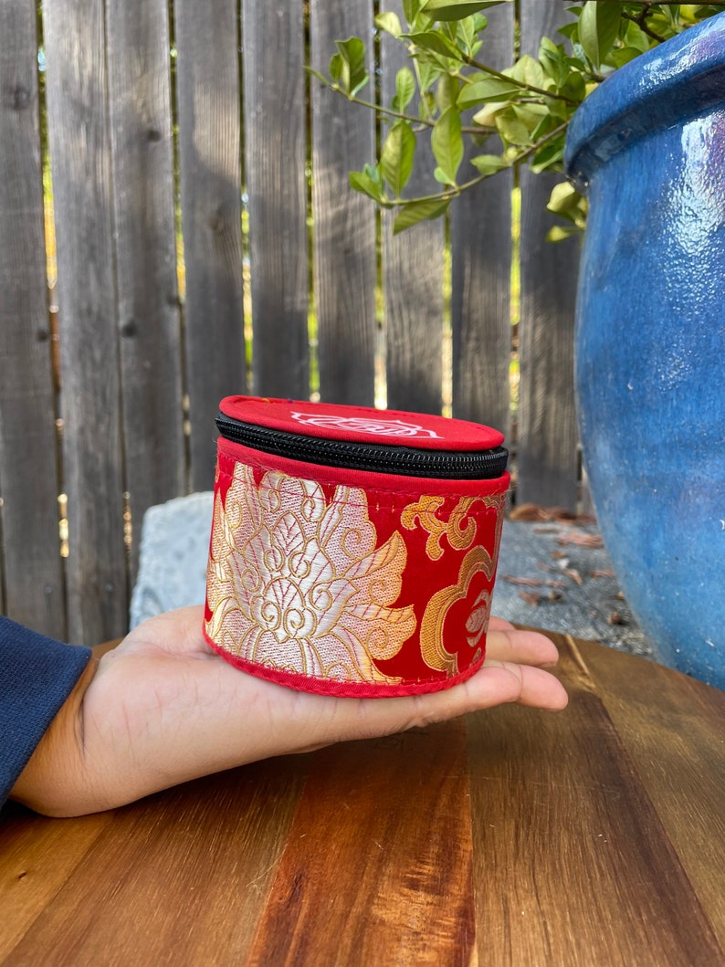 May include: A small, round, red and gold fabric box with a black zipper. The box has a floral pattern and is being held in a hand.